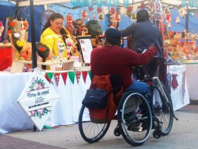 En la imagen se ve desde atrás, a un hombre en silla de ruedas que está observando un stand de un emprendomiento que vende flores de papel, la cual es atendida por una mujer.